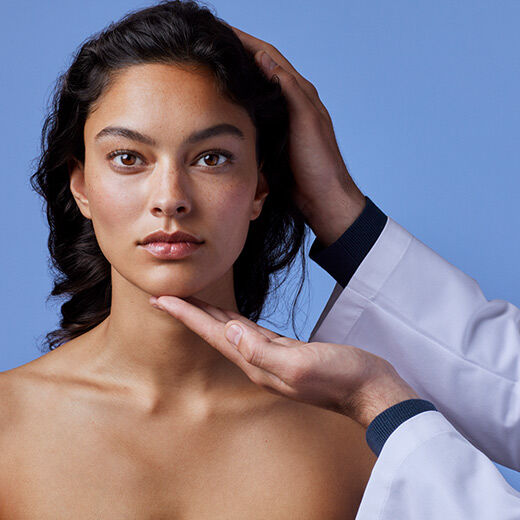 A close-up of a woman with long, dark hair and natural makeup, gazing directly at the camera. A hand in a white coat gently supports her chin against a soft blue background.