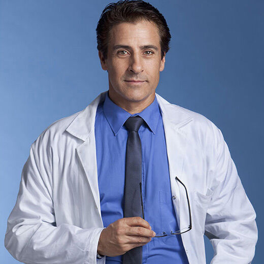A confident male doctor in a white lab coat and blue shirt, smiling against a blue background.