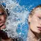 A close-up of two models with different skin tones, surrounded by splashing water against a bright blue background. The mood is fresh and summery.