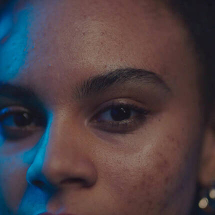 Close-up of a person's face, featuring dark hair, glowing skin, and intense eyes illuminated by blue lighting.