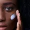 A close-up of a person with curly hair applying a skincare product to their cheek against a blue background.