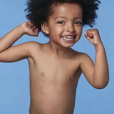 A joyful child with curly hair smiles widely, showcasing their teeth against a light blue background.