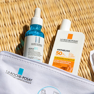 A close-up of two skincare products from La Roche-Posay: a blue bottle of Hyalu B5 Serum and a white tube of Anthelios 50+ sunscreen, placed on a textured surface.