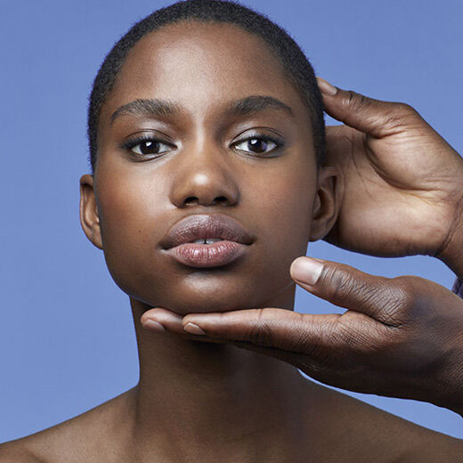 A close-up of a person’s face with smooth skin and natural makeup, while a hand gently adjusts their chin against a blue background.