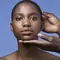A close-up portrait of a person with short hair, showcasing a hand gently touching their face against a blue background.