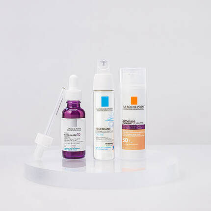 Three skincare products displayed on a circular pedestal: a dark purple serum with a dropper, a white moisturizer in a pump bottle, and a light orange sunscreen in a cream tube. All products are from the La Roche-Posay brand.