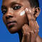 A close-up of a person applying a skincare product to their cheek against a blue background.