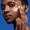 A close-up of a person applying a skincare product to their cheek against a blue background.