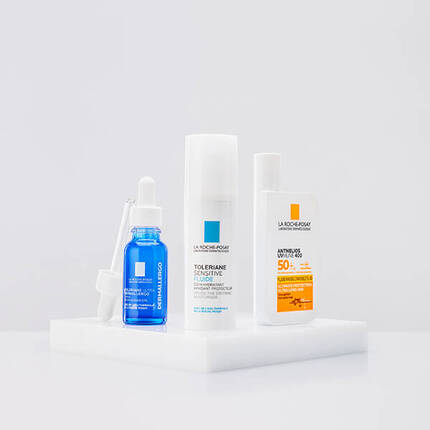 A collection of three La Roche-Posay skincare products displayed on a white pedestal. The products include a blue serum with a dropper, a white fluid moisturizer, and a sunscreen with SPF 50.
