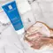 A hand lathering a foaming facial cleanser on a marble surface, with a towel and the cleanser's bottle visible in the background.