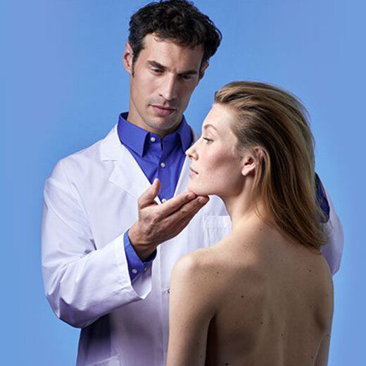 A healthcare professional examines a woman's face, both appearing focused and engaged, against a blue background. The woman has her eyes closed and looks serene.