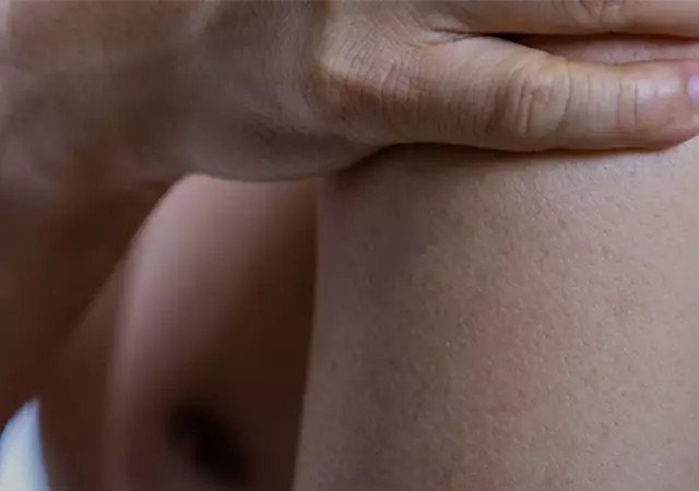 A close-up of a person's bare shoulder and arm, with one hand gently resting on the shoulder. The skin appears smooth and the background is softly blurred.