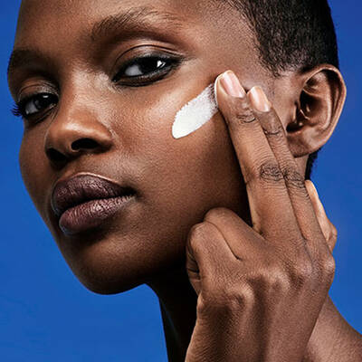 A woman applies a skincare product to her face while looking directly at the camera, against a blue background.