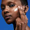 A woman applies a skincare product to her face while looking directly at the camera, against a blue background.