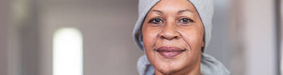 A woman with a warm smile, wearing a gray headscarf and a simple top, stands in a softly lit indoor setting.