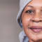 A woman with a warm smile, wearing a gray headscarf and a simple top, stands in a softly lit indoor setting.