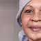 A woman with a warm smile, wearing a gray headscarf and a simple top, stands in a softly lit indoor setting.