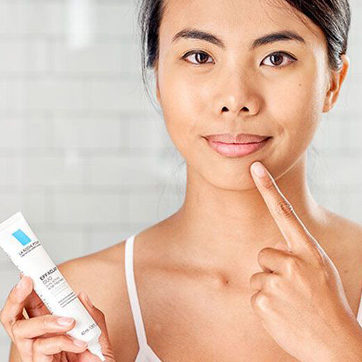 A person with a light skin tone is pointing to their chin while holding a tube of skincare product in a bright bathroom setting.