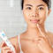 A person with a light skin tone is pointing to their chin while holding a tube of skincare product in a bright bathroom setting.