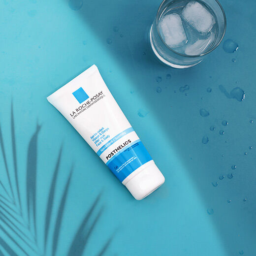 A tube of La Roche-Posay Posthelios after-sun care lotion on a blue background, with water droplets and a shadow of a plant in the foreground.