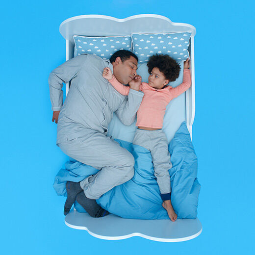 A father and his young child are lying side by side in bed, both looking relaxed. The child, with curly hair, is playfully poking the father's face. The bedding features a light blue background with white clouds.