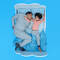A father and his young child are lying side by side in bed, both looking relaxed. The child, with curly hair, is playfully poking the father's face. The bedding features a light blue background with white clouds.