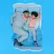 A father and his young child are lying side by side in bed, both looking relaxed. The child, with curly hair, is playfully poking the father's face. The bedding features a light blue background with white clouds.