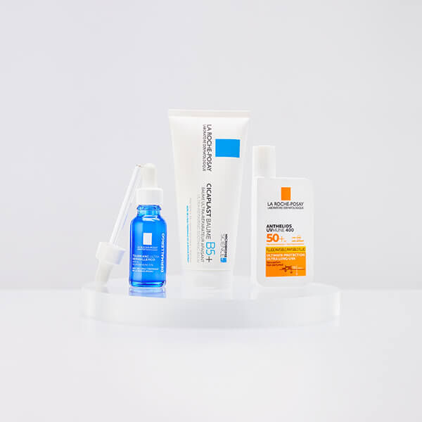 Four skincare products displayed on a clear stand, including a blue serum, a white cream tube, and two sunscreen bottles with orange and white packaging.
