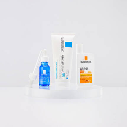 Four skincare products displayed on a clear stand, including a blue serum, a white cream tube, and two sunscreen bottles with orange and white packaging.