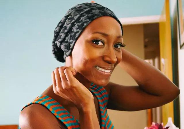 A smiling woman with a patterned headscarf adjusts her hair, set against a light blue background.