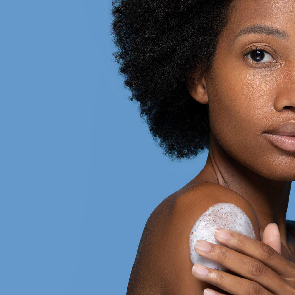 A woman with natural curly hair applies a skincare product to her shoulder, set against a solid blue background.