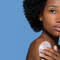 A woman with natural curly hair applies a skincare product to her shoulder, set against a solid blue background.