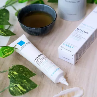 A tube of La Roche-Posay Effaclar Duo+ anti-blemish cream next to its box, placed on a wooden surface with a green plant nearby.