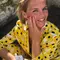 A smiling person with short blonde hair and colorful nails, wearing a yellow polka dot shirt and gently touching their face.