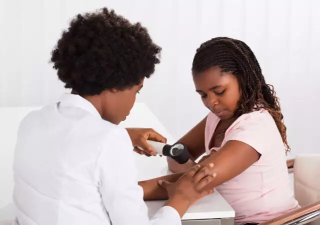 A young girl in a pink shirt is sitting while a caregiver checks her blood pressure with a monitor.
