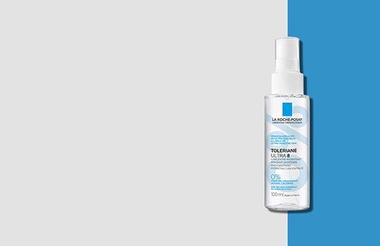 A clear spray bottle of a hair treatment product on a split background of light gray and bright blue.