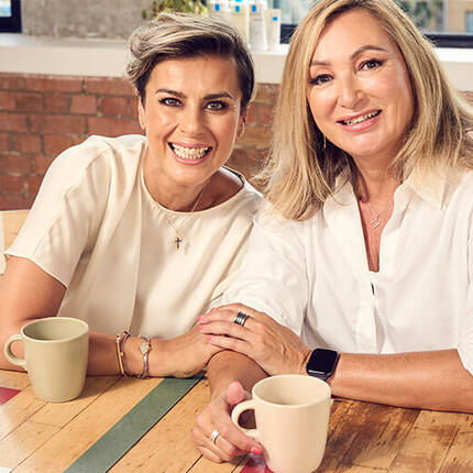 Two women with cheerful expressions are sitting together, smiling at the camera. They have stylish hairstyles and are in a bright, inviting indoor setting with a glimpse of skincare products in the background.