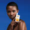 A woman holds a bottle of La Roche-Posay Anthelios sunscreen in front of her face, against a blue background. She has a confident expression, showcasing her skin.