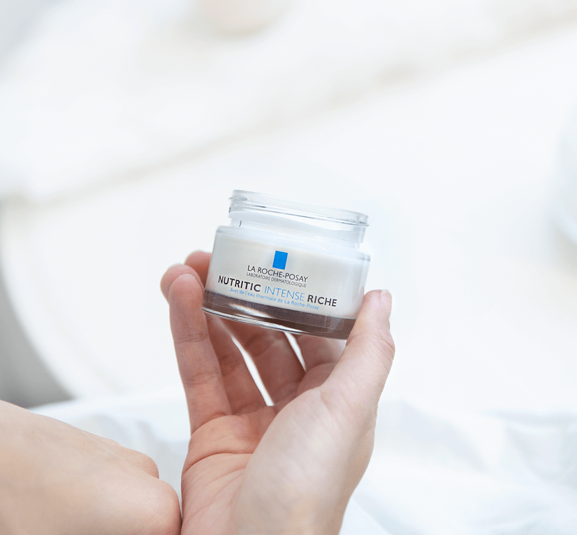 A hand holding a jar of La Roche-Posay Nutritic Intense Riche cream against a soft, blurred background.