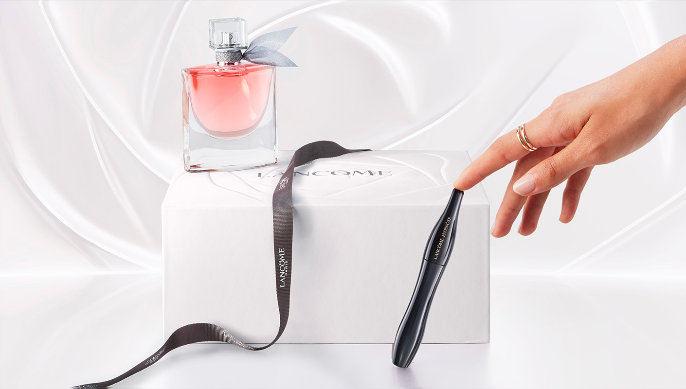 A hand reaching towards a Lancôme perfume bottle and mascara on a white box with a ribbon, showcasing luxury beauty products.