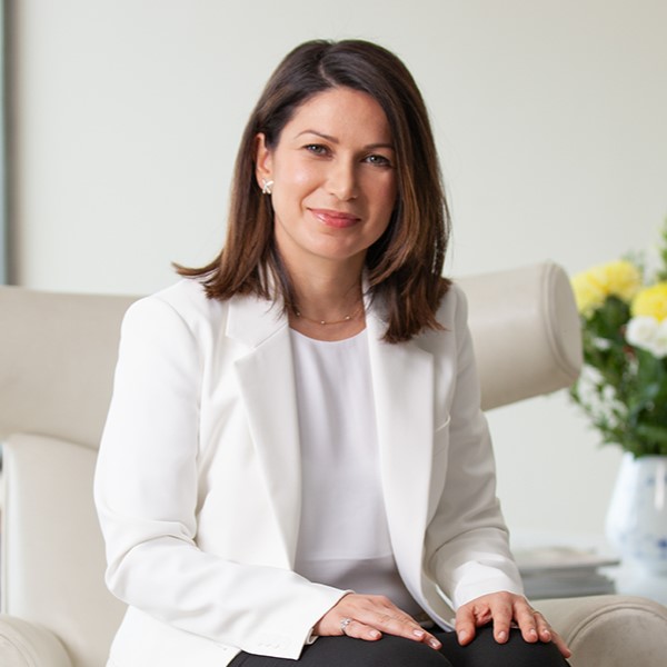 Two bottles of Anthelios UVMUNE 400 Invisible Fluid SPF50+, with one positioned slightly behind the other against a blue background. Dr. Hiva Fassihi sitting on a chair in a clinic