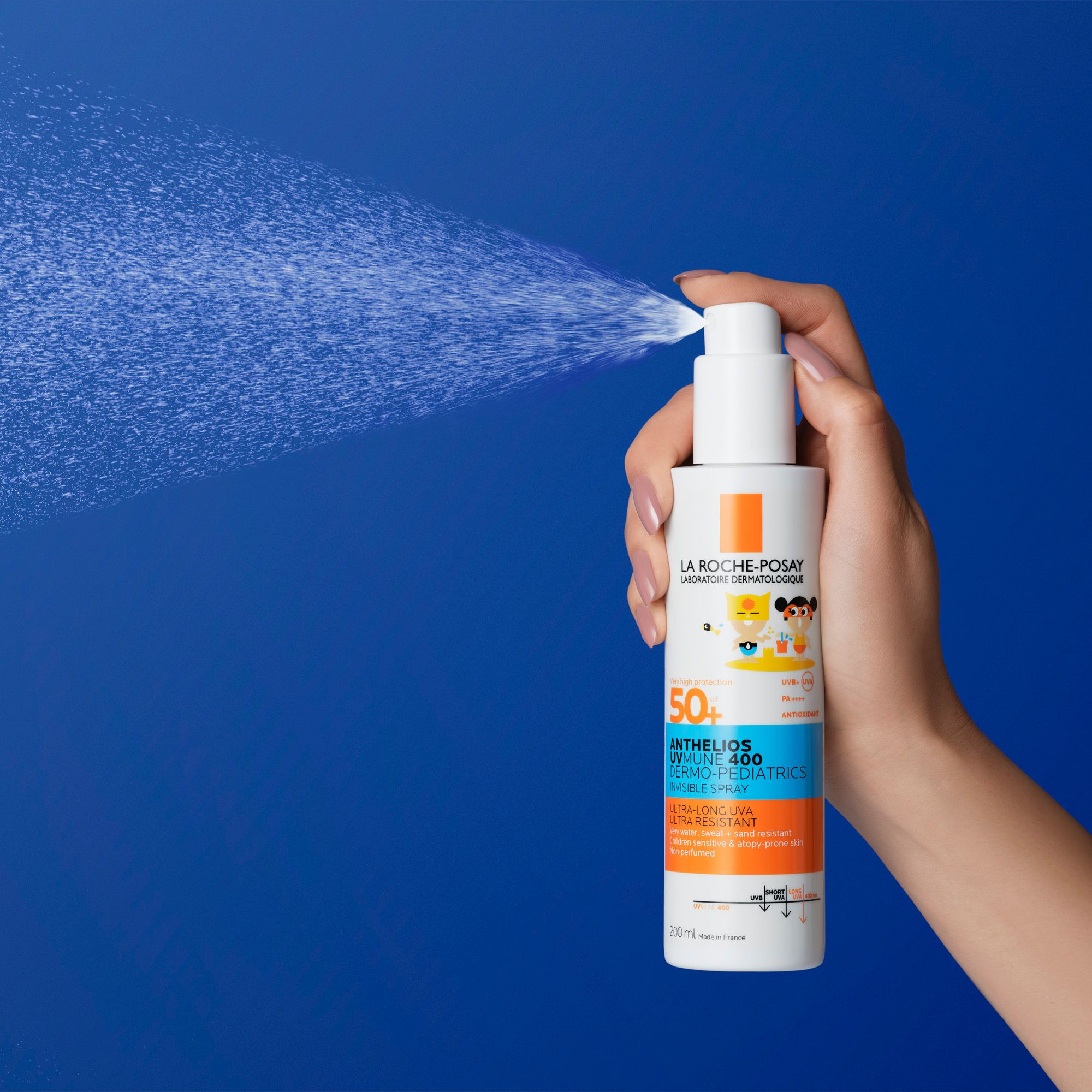 A hand pumps the nozzle of a bottle of Anthelios Invisible Kids Spray SPF50+ to release a fine mist of sunscreen, highlighting its easy, spray-on application for children.